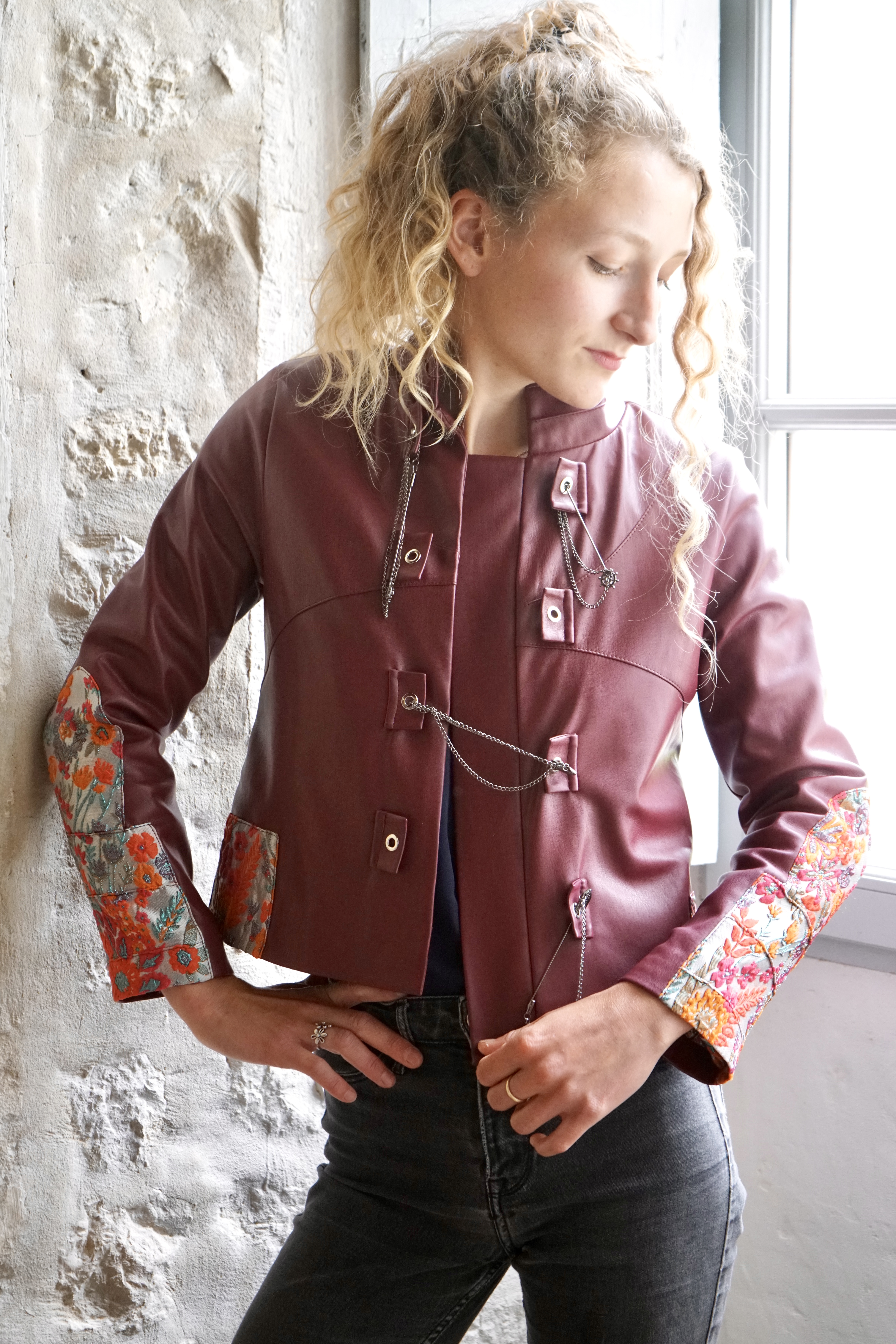 Model wearing a bespoke tailored vegan leather jacket in deep red-brown with embroidered orange-red floral patches, Mongolian warrior inspired design, unique metal clasps, luxurious lining matching the external embroidered patches 